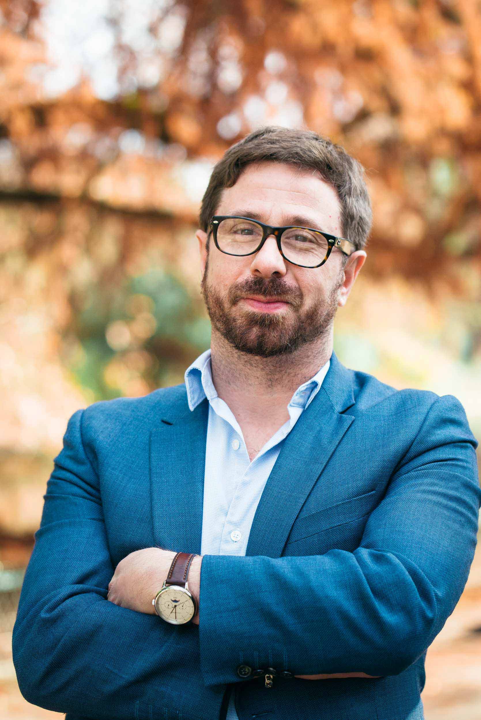 Un homme barbu aux cheveux courts et portant des lunettes se tient debout, les bras croisés, avec assurance, dégageant une ambiance de portraitiste professionnel. Il porte un blazer bleu sur une chemise bleu clair et une montre-bracelet. L'arrière-plan présente un feuillage automnal flou dans des tons orange et marron, rappelant Bordeaux, en Gironde.