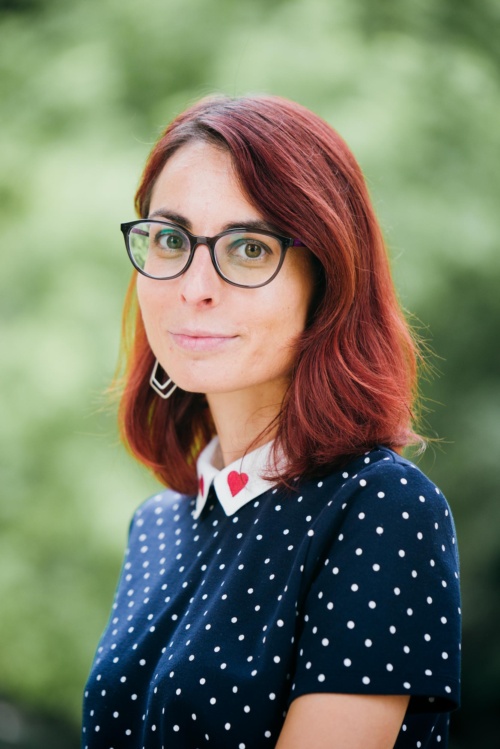 Une femme aux cheveux roux mi-longs, portant des lunettes à monture noire, pose en extérieur dans un portrait d'entreprise. Elle porte un haut à pois bleu marine avec un col blanc orné d'un petit cœur rouge. L'arrière-plan est une verdure floue, la mettant en valeur comme le sujet principal de la photo prise à Bordeaux, en Gironde.