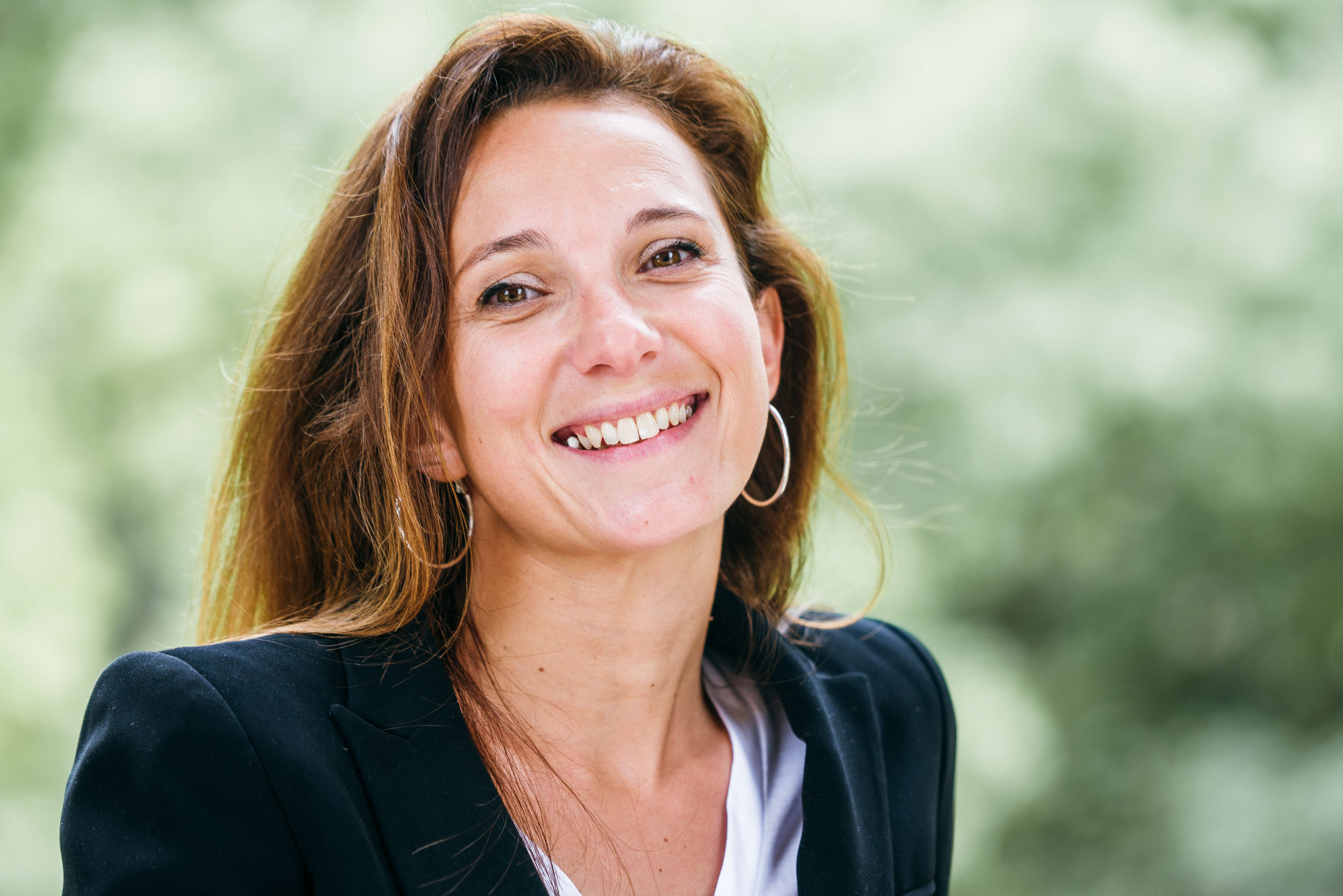 Une femme aux longs cheveux bruns sourit chaleureusement à l'objectif. Elle porte un blazer noir sur un haut blanc et des créoles. L'arrière-plan, de couleur bordeaux, présente un feuillage vert flou qui donne une sensation douce et naturelle à ce portrait d'entreprise professionnel.