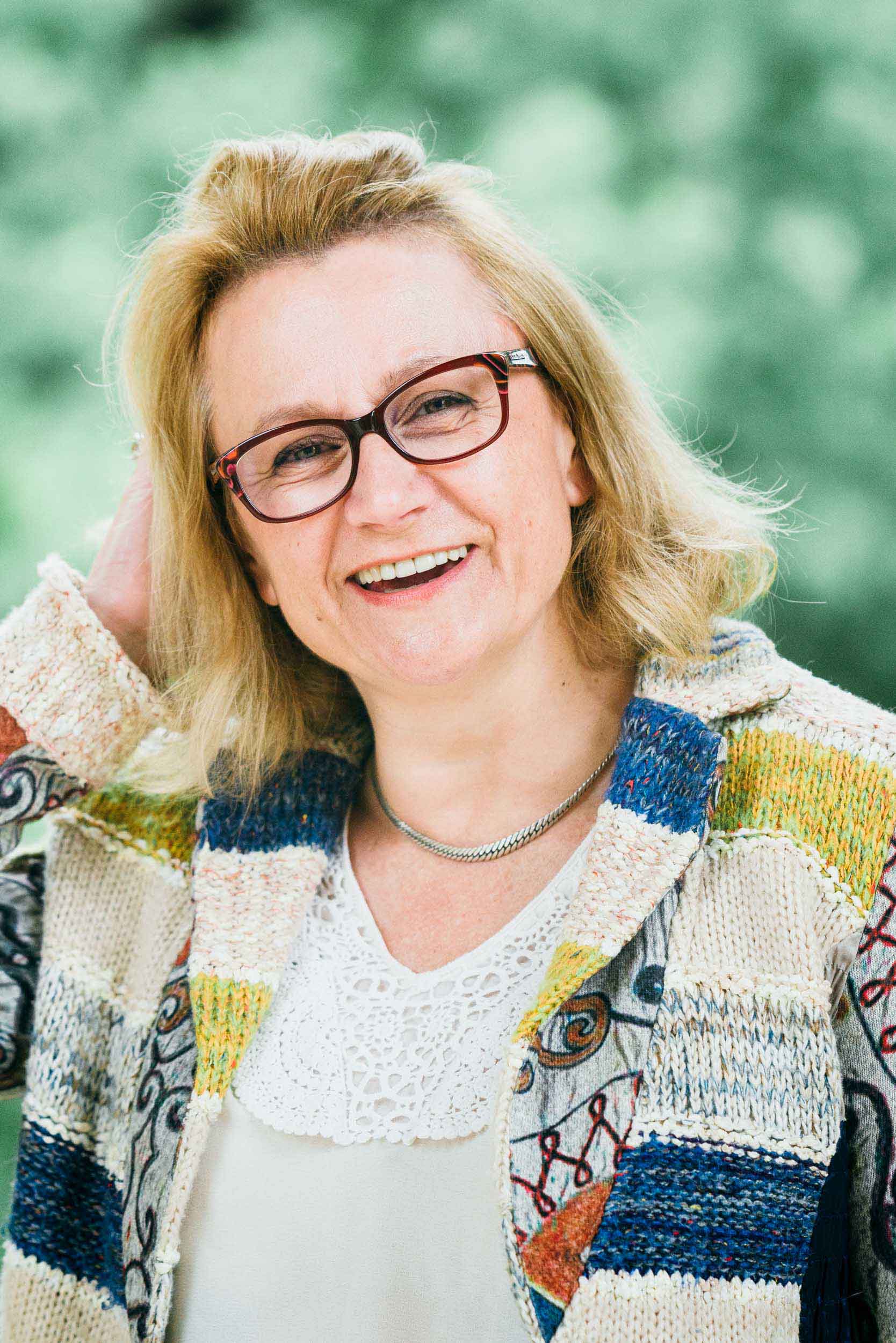 Une femme souriante aux cheveux blonds mi-longs et portant des lunettes se tient à l'extérieur. Elle porte un cardigan coloré à motifs sur un chemisier blanc avec des détails en dentelle. Elle a une main levée pour toucher ses cheveux. Le fond vert flou suggère un feuillage, idéal pour un portrait d'entreprise à Bordeaux ou en Gironde.