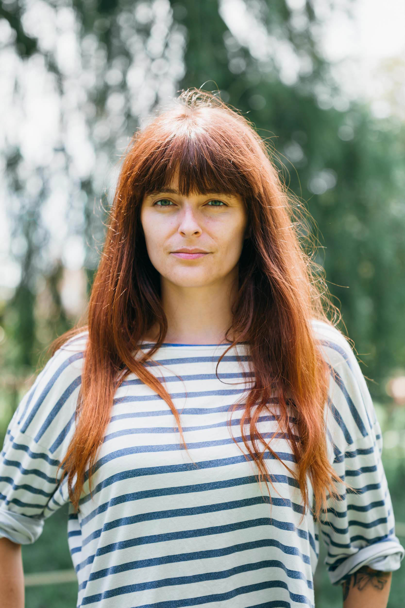 Une femme aux cheveux longs et raides de couleur auburn se tient debout dans un décor extérieur à Bordeaux. Elle porte une chemise rayée à manches longues et a une expression neutre. Derrière elle, on voit des arbres et des feuillages verts flous, suggérant un environnement naturel. L'éclairage est vif, indiquant une journée ensoleillée.