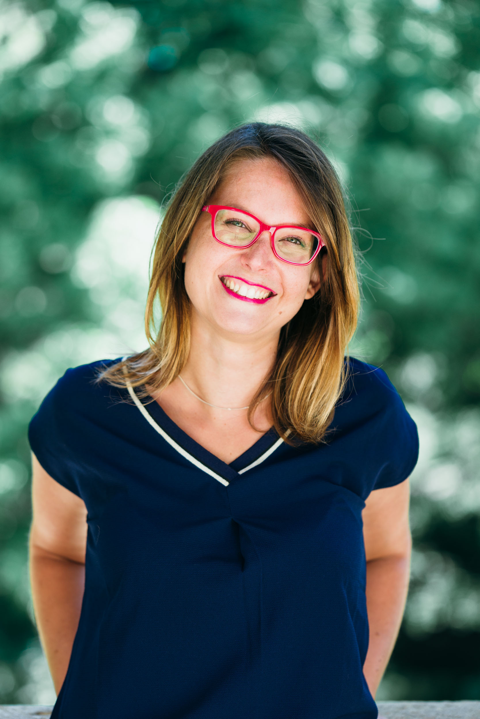 Une personne souriante aux cheveux châtains mi-longs porte des lunettes rouge vif et un haut bleu foncé à décolleté en V. Ses mains sont derrière son dos et l'arrière-plan présente un effet bokeh vert et blanc flou, suggérant un décor extérieur, parfait pour un portrait d'entreprise à Bordeaux.