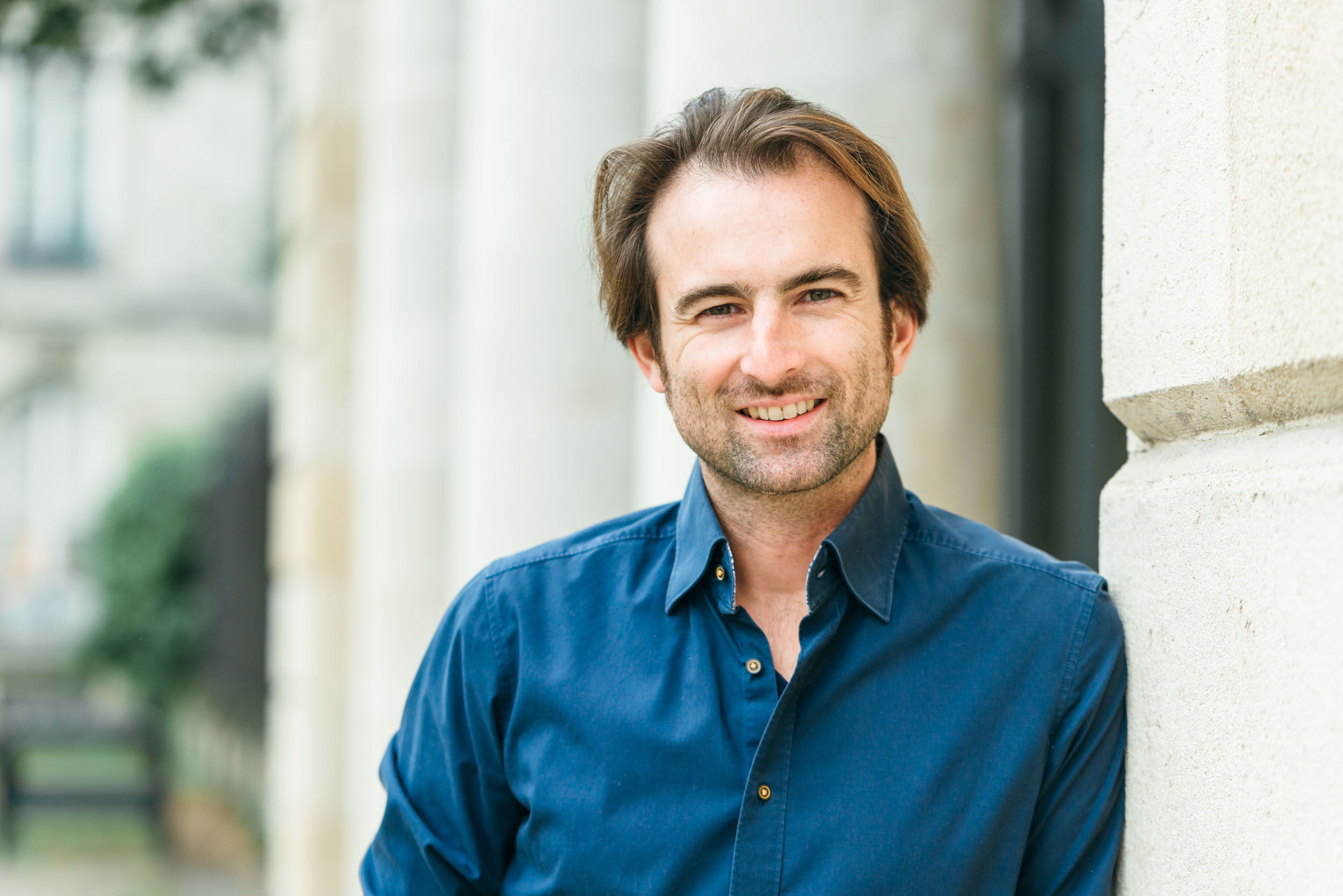 Un homme aux cheveux châtains mi-longs et à la barbe claire sourit en se tenant à l'extérieur devant un bâtiment aux colonnes de pierre, incarnant l'essence d'un portrait d'entreprise. Il porte une chemise bleu foncé et semble détendu, avec un arrière-plan flou montrant de la verdure et une partie du bâtiment à Bordeaux, en Gironde.