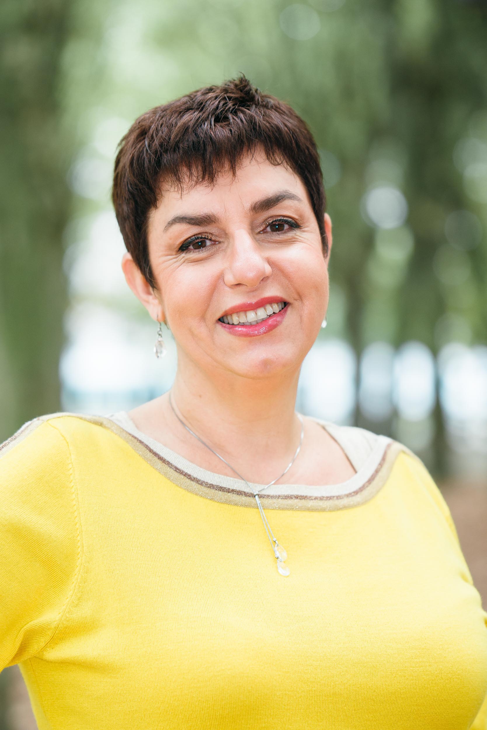 Une femme aux cheveux bruns courts sourit chaleureusement à l'objectif. Elle porte un haut jaune vif avec un décolleté beige et un délicat collier argenté, parfait pour un portraitiste professionnel. L'arrière-plan est flou mais présente un feuillage vert, suggérant un décor naturel en plein air à Bordeaux.