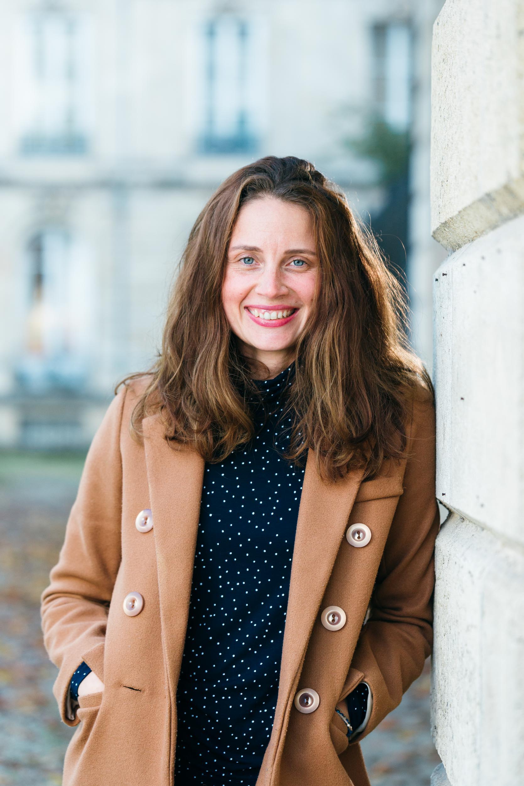 Une femme aux longs cheveux bruns sourit en se tenant dehors devant un bâtiment flou à Bordeaux. Elle porte un manteau beige sur une tenue bleu marine à pois blancs, les mains confortablement dans les poches de son manteau. Ses cheveux sont détachés et le paysage urbain derrière elle met en valeur l'architecture historique de la Gironde.