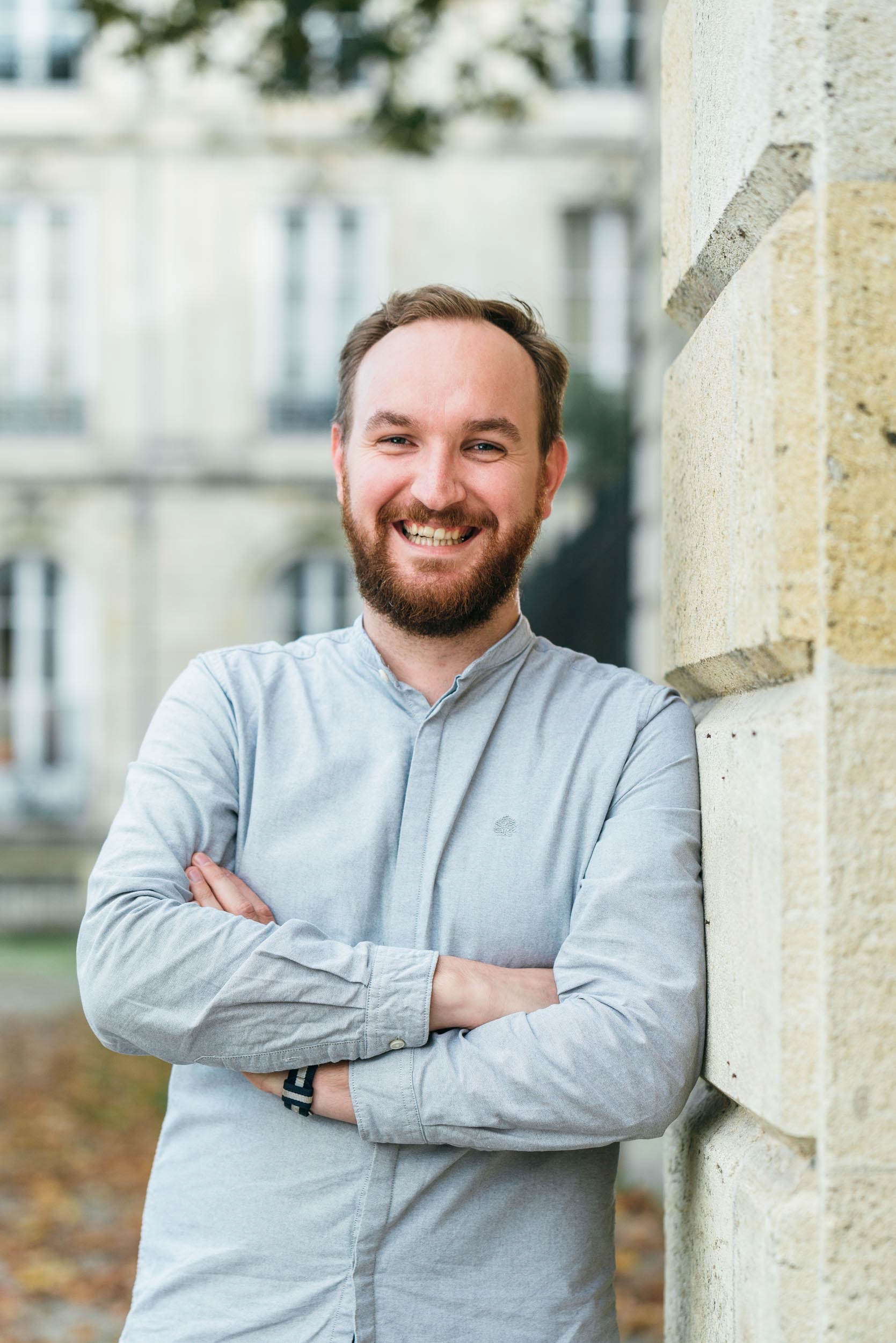 Un homme barbu est appuyé contre un mur de pierre en plein air à Bordeaux, souriant, les bras croisés. Vêtu d'une chemise gris clair et portant une montre au poignet gauche, il dégage une atmosphère confiante et professionnelle. L'arrière-plan flou présente des fenêtres et des feuilles d'automne éparpillées sur le sol.