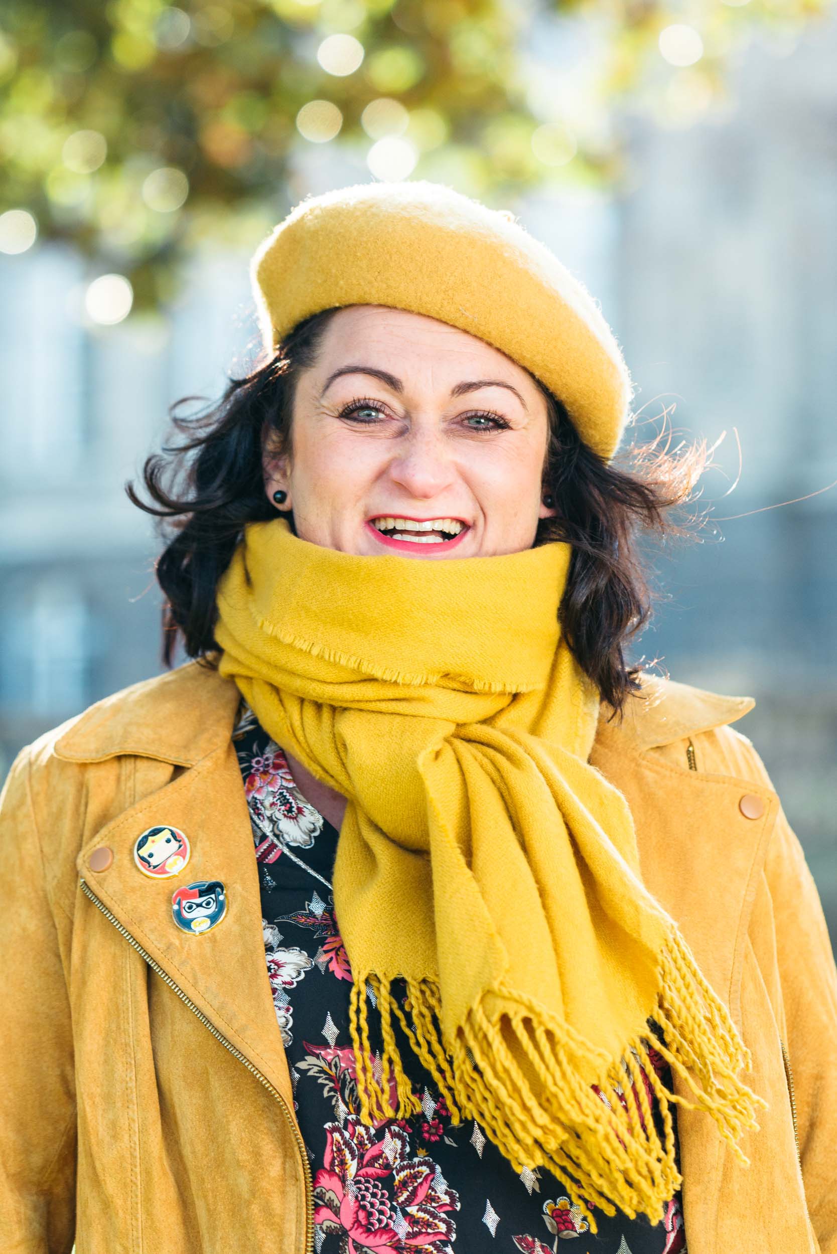 Une femme sourit avec éclat dans un décor extérieur, portant un béret jaune, un foulard jaune assorti et une veste couleur moutarde ornée de broches en émail. Son haut fleuri ajoute une touche de charme. L'arrière-plan flou suggère la verdure et les bâtiments d'une journée ensoleillée à Bordeaux, créant le portrait professionnel parfait.