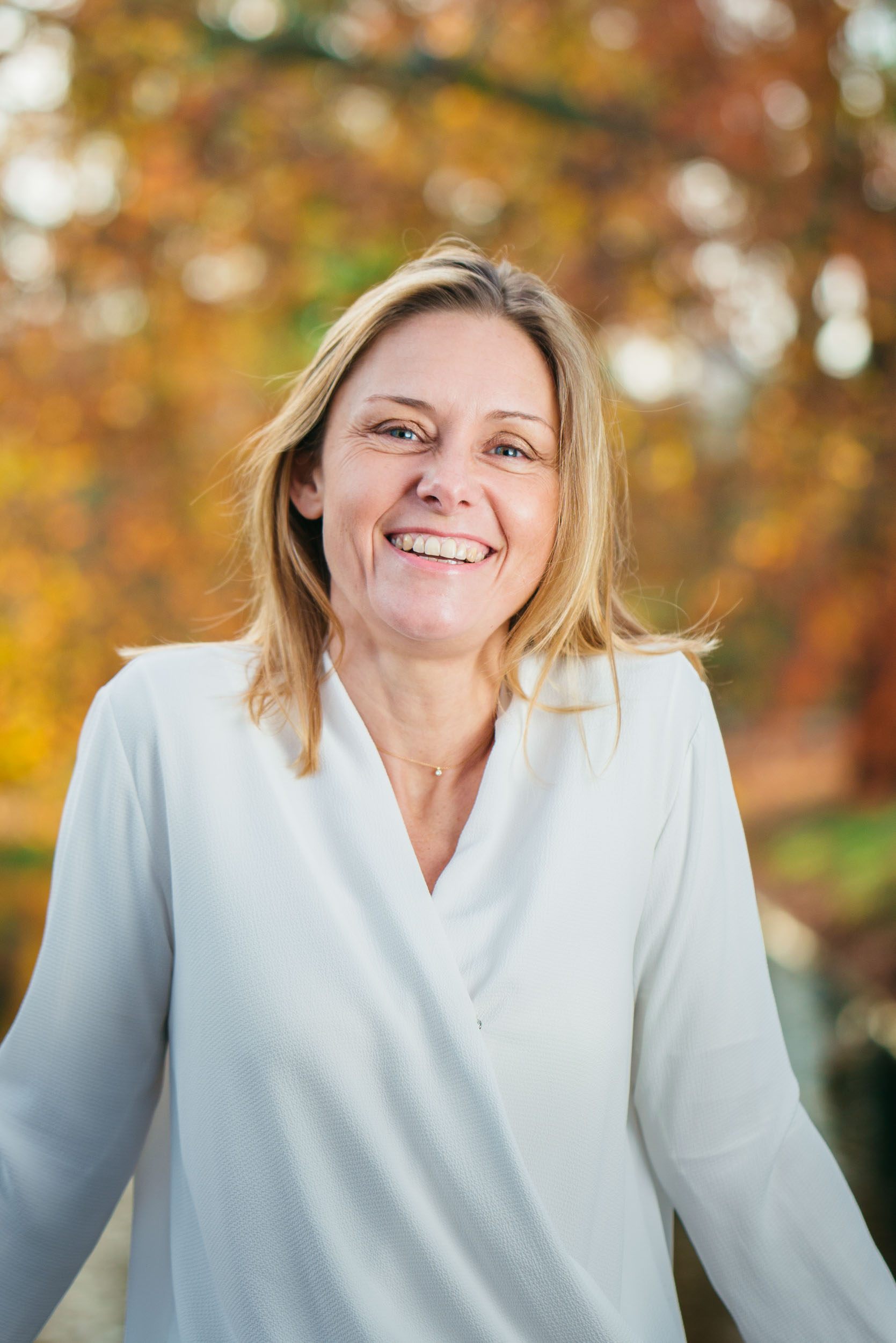 Une femme souriante aux cheveux blonds mi-longs se tient devant un arrière-plan automnal flou à Bordeaux, en Gironde. Elle porte un chemisier blanc à col en V et semble détendue et heureuse. Le portrait professionnel capture magnifiquement les teintes chaudes du feuillage d'automne orange et jaune.