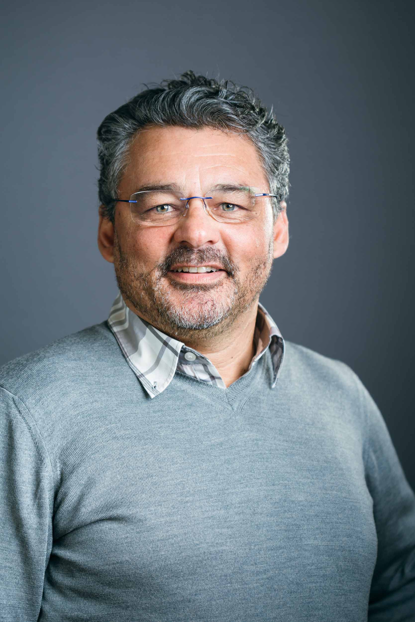 Un homme d'âge moyen aux cheveux courts, bouclés, poivre et sel, et portant une barbe sourit à l'objectif. Il porte des lunettes, un pull gris sur une chemise à col et se tient debout sur un fond sombre et uni. Sa tenue et son comportement évoquent un portrait d'entreprise à Bordeaux ou en Gironde.