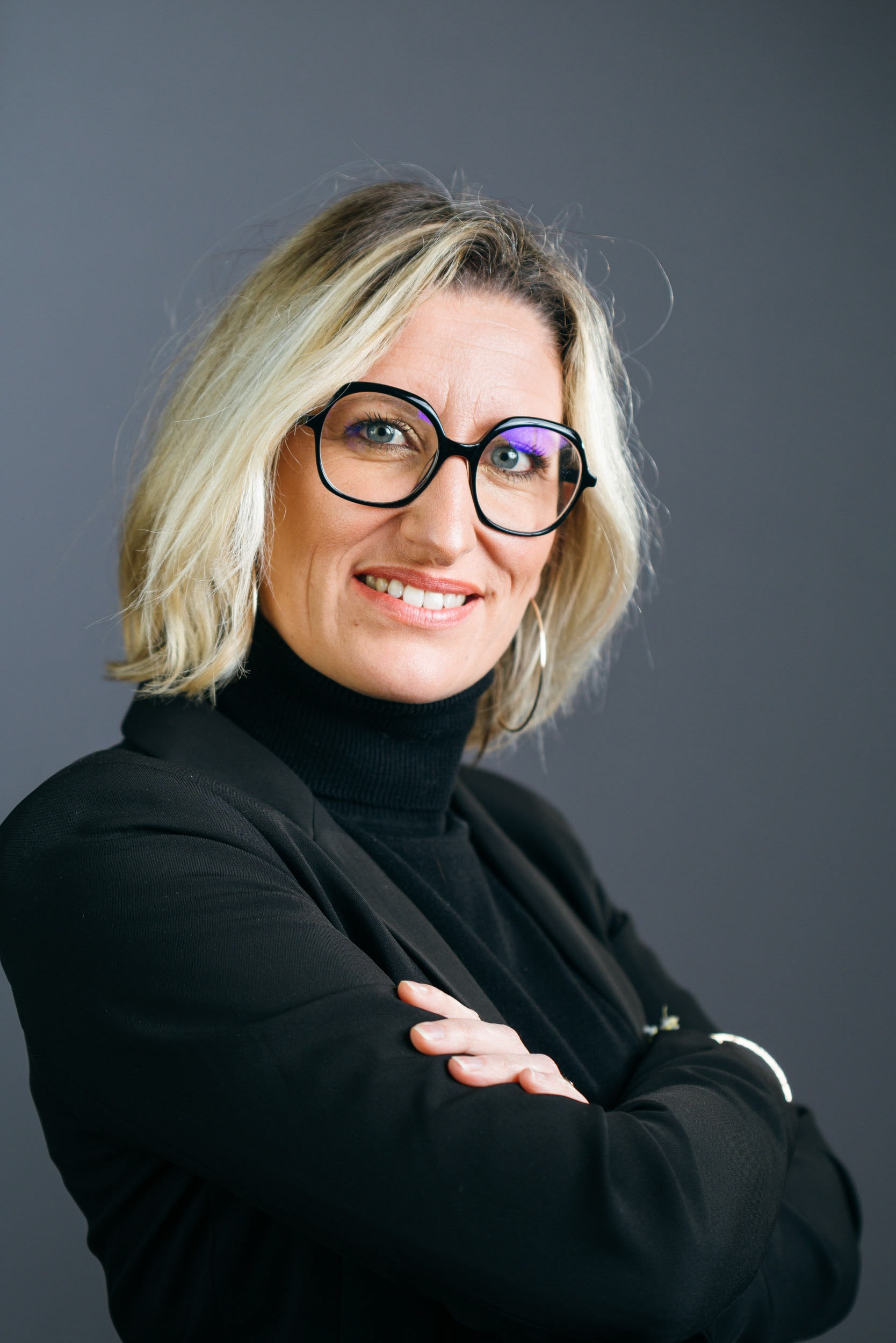 Une femme aux cheveux blonds mi-longs et aux lunettes rectangulaires noires sourit avec assurance sur ce portrait d'entreprise. Elle porte un col roulé noir et un blazer noir, les bras croisés. Le fond gris uni met en valeur son attitude professionnelle, parfaite pour une utilisation dans des environnements bordelais ou girondins.