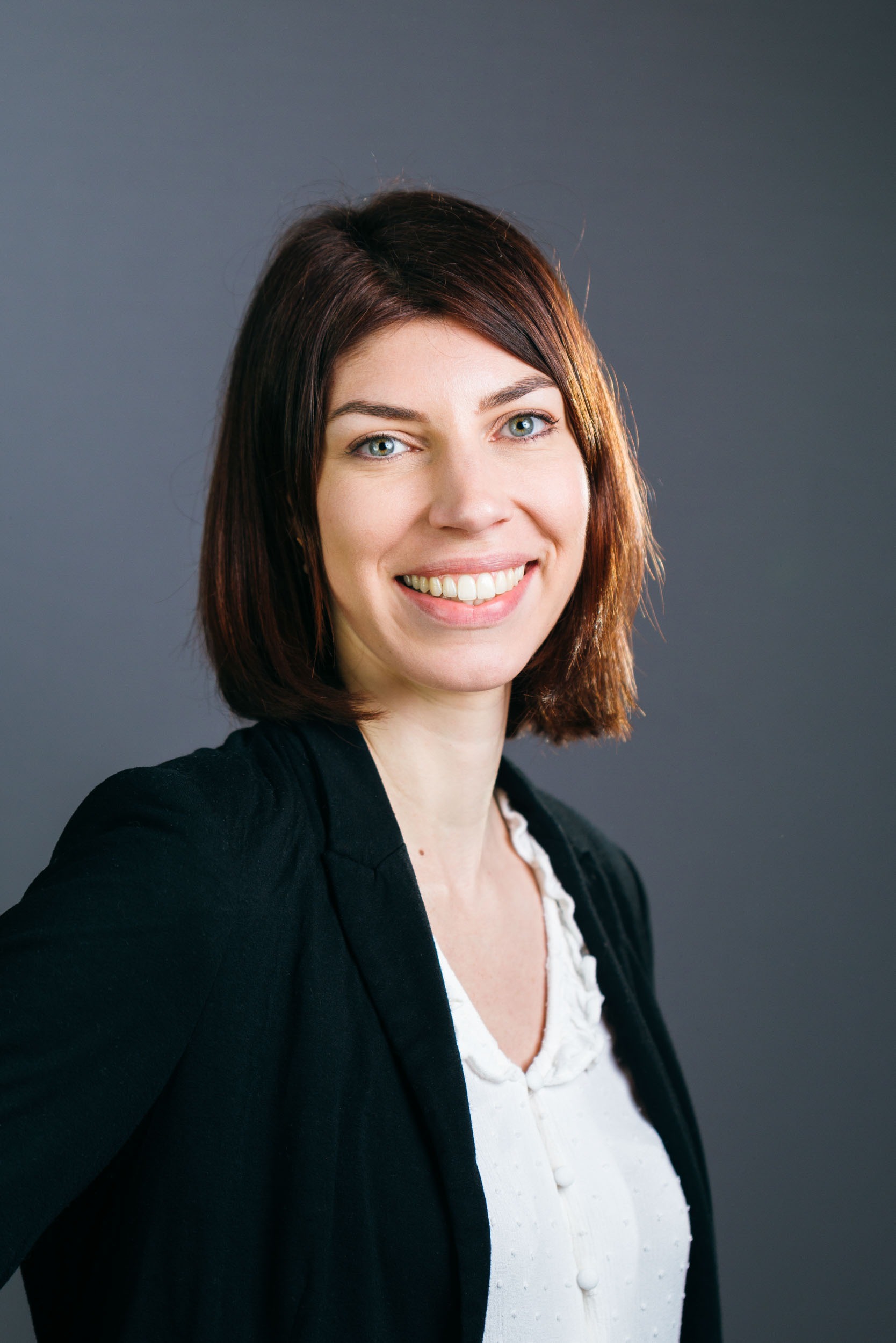 Une femme aux cheveux bruns raides et longs sourit chaleureusement à l'objectif dans ce portrait d'entreprise. Elle porte un chemisier blanc sous un blazer noir. Le fond gris uni accentue son expression amicale et sa tenue professionnelle, créant un look soigné idéal pour tout environnement d'affaires à Bordeaux.