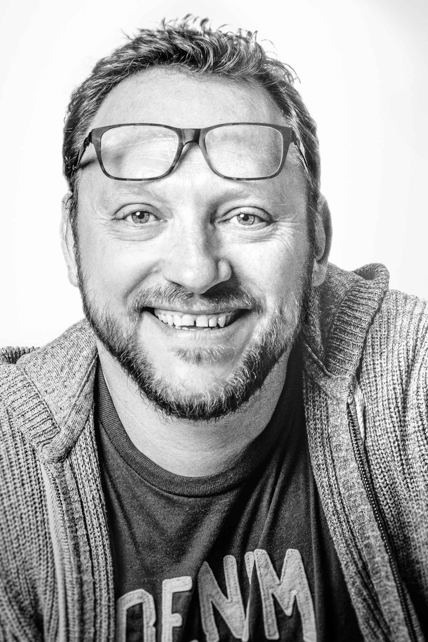 Image en noir et blanc d'un homme souriant aux cheveux courts et à la barbe, portant des lunettes perchées sur son front. Vêtu d'un pull décontracté et d'un t-shirt, il respire la chaleur. L'arrière-plan uni attire l'attention sur son expression joyeuse, parfaitement capturée par un photographe pour les entreprises.