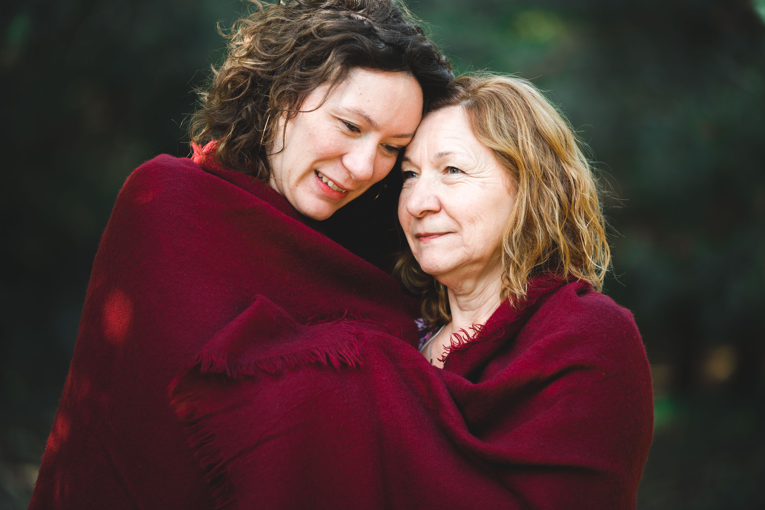 Deux femmes, représentées dans une séance photo réconfortante, mère-fille, sont enveloppées ensemble dans une couverture rouge foncé, partageant un moment affectueux. La plus jeune femme, aux cheveux noirs bouclés, sourit et regarde vers le bas, tandis que la femme plus âgée aux cheveux clairs ondulés pose sa tête sur l'épaule de la plus jeune sur un arrière-plan extérieur flou.