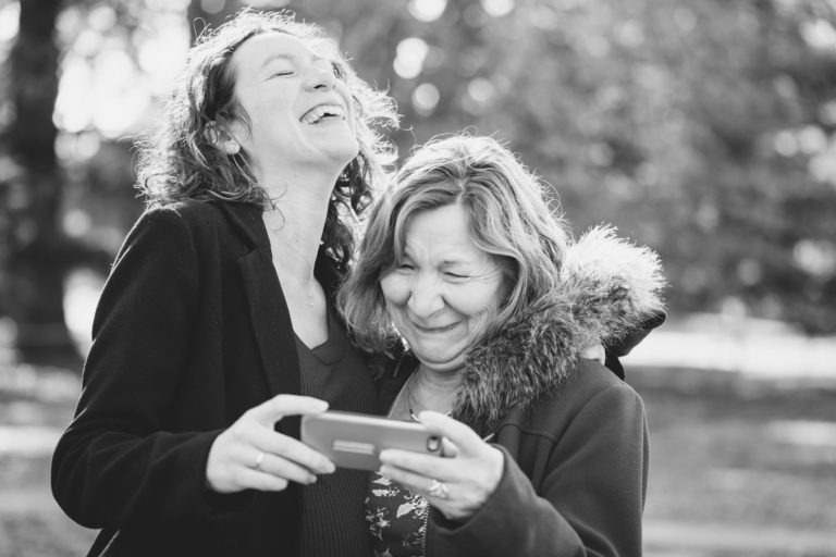 Deux femmes se tiennent dehors, souriant et riant ensemble dans ce qui semble être une charmante séance photo mère-fille. Une femme, aux cheveux bouclés, se penche vers l'autre, son bras autour de ses épaules. L'autre femme, aux cheveux ondulés et portant une veste doublée de fourrure, tient un smartphone sur lequel apparaît quelque chose d'amusant. L'arrière-plan est flou et naturel.