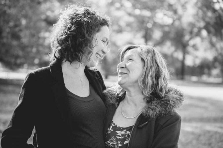 Une photo en noir et blanc montre une mère et sa fille à l'extérieur, debout près l'une de l'autre. Toutes deux sourient et se regardent dans les yeux. La femme de gauche a les cheveux courts et bouclés et porte une veste sombre par-dessus un haut. La femme de droite a les cheveux ondulés et porte un manteau avec un col doublé de fourrure. L'arrière-plan est flou, montrant des arbres, capturant l'essence de