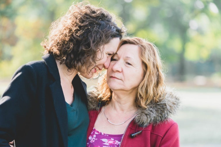 Deux femmes se tiennent côte à côte à l'extérieur. La femme de gauche, aux cheveux bruns bouclés, sourit et touche son front à celui de la femme de droite, qui a les cheveux blonds ondulés et une expression pensive. Sur cette photo de séance de spiritisme mère-fille, la femme de droite porte un manteau rouge avec une capuche doublée de fourrure et un haut à fleurs roses.