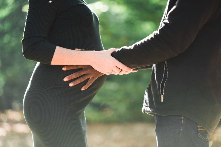 Gros plan d'une femme enceinte en robe noire et d'une personne en veste noire debout à l'extérieur. La personne tient doucement le ventre de la femme d'une main et sa main de l'autre. La lumière du soleil filtre à travers le feuillage vert, créant une atmosphère sereine parfaite pour cette séance photo mère-fille.