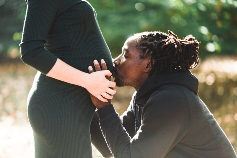 Un homme aux dreadlocks embrasse le ventre rond d'une femme enceinte vêtue d'une robe vert foncé. La femme est debout et l'homme est à genoux devant elle dans un décor extérieur rappelant une séance photo tendre entre mère et fille. La lumière du soleil filtre à travers la verdure en arrière-plan.