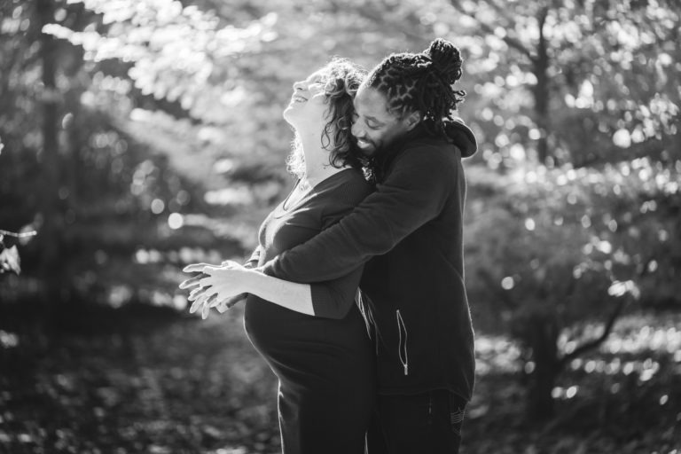 Une photographie en noir et blanc évoque la tendresse d'une séance photo mère-fille, mettant en scène une femme enceinte et un homme enlacés dans un espace boisé serein. La femme, souriante et berçant son ventre, se tient devant tandis que l'homme la serre dans ses bras par derrière, posant ses mains sur les siennes. La lumière du soleil filtre à travers les arbres, projetant une douce lueur autour d'eux.