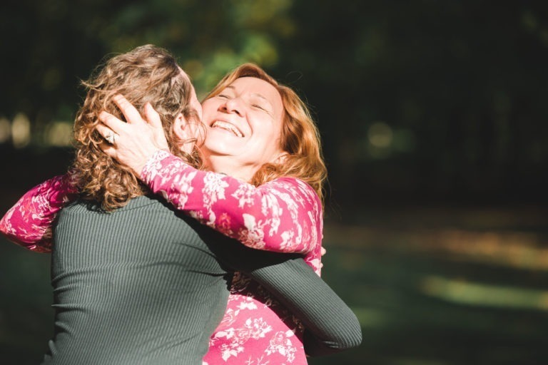 Deux personnes s'embrassent chaleureusement dans un parc ensoleillé lors d'une séance photo mère-fille. L'une des personnes, aux cheveux bouclés, porte un haut vert foncé et se détourne de l'objectif. L'autre personne, aux cheveux mi-longs ondulés dans un haut à fleurs roses, sourit radieusement, les yeux fermés alors qu'elles s'embrassent au milieu d'une verdure floue.