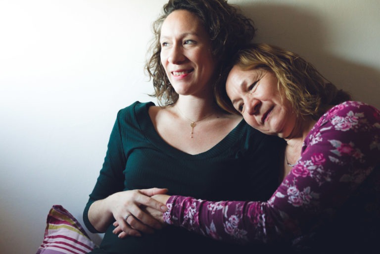 Deux femmes sont assises l'une à côté de l'autre. Celle de gauche, aux cheveux bouclés et vêtue d'un haut vert foncé, sourit doucement tandis que celle de droite, vêtue d'un haut rouge à motifs floraux, pose sa tête sur l'épaule de l'autre. Leur pose intime pourrait facilement être capturée dans une séance photo mère-fille.