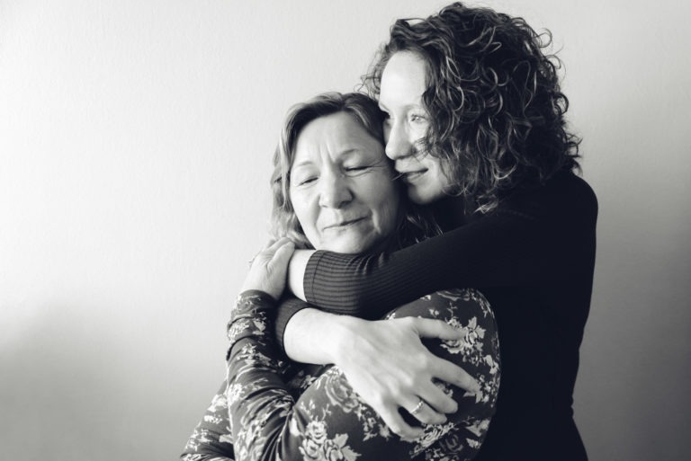 Une photo en niveaux de gris montre deux femmes, peut-être dans une séance photo mère-fille, s'embrassant affectueusement. La femme la plus âgée ferme les yeux, posant sa tête sur l'épaule de la plus jeune, qui sourit doucement les yeux ouverts. Toutes deux ont les cheveux bouclés. Le fond mural uni et clair met en valeur leur lien chaleureux et solidaire.