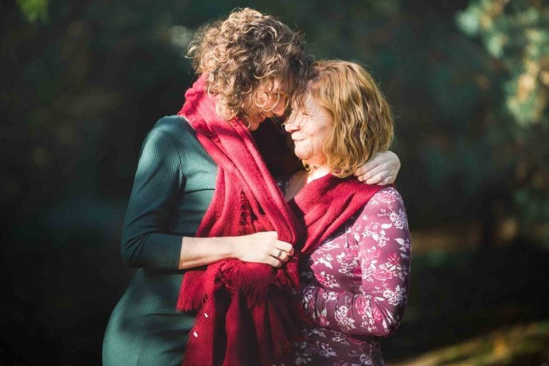 Deux personnes se tiennent côte à côte à l'extérieur, toutes deux portant un foulard rouge. La personne de gauche a les cheveux bouclés, une robe verte et passe son bras autour de l'autre personne. Sur cette touchante séance photo mère-fille, elles partagent un moment affectueux, se touchant le front et souriant.