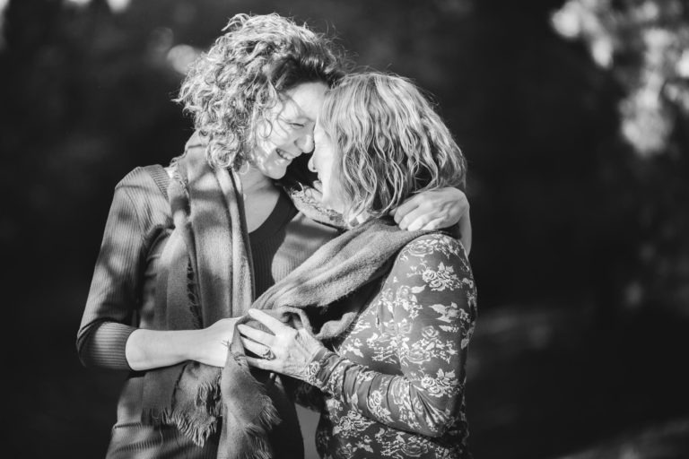 Une photo en noir et blanc d'une mère et de sa fille debout côte à côte, face à face, leurs têtes se touchant. Toutes deux sourient chaleureusement, l'une a son bras enroulé autour des épaules de l'autre. Elles semblent vivre un moment d'affection et de connexion, entourées d'un décor extérieur flou et naturel - une séance photo mère-fille parfaite.