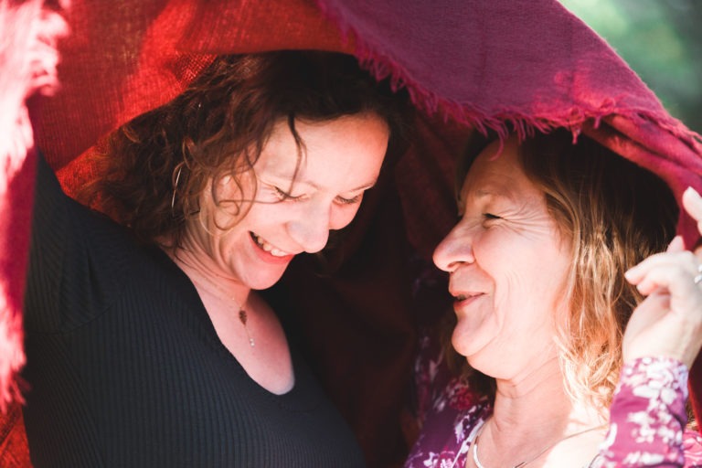 Deux femmes partagent un moment de légèreté sous une couverture rouge drapée. La femme de gauche, aux cheveux bruns bouclés et au haut noir, rit, tandis que la femme de droite, aux cheveux blonds ondulés et au chemisier à motifs, la regarde avec affection. La lumière du soleil filtre à travers la couverture dans cette séance photo réconfortante entre mère et fille.