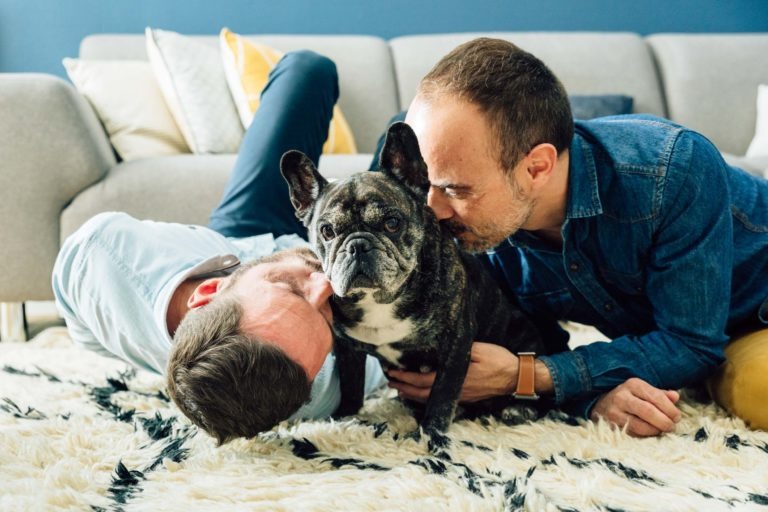 Deux personnes allongées sur un tapis blanc texturé, face à un chien noir et blanc aux grandes oreilles. Une personne, allongée sur le côté, la tête posée sur le tapis, sourit au chien. L'autre personne, accroupie, embrasse la tête du chien dans un geste d'amour inconditionnel. Un canapé gris avec des coussins à motifs est visible en arrière-plan.