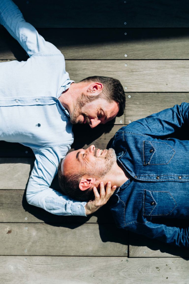 Deux hommes sont allongés sur le dos sur une terrasse en bois, les têtes rapprochées et se souriant. L'un porte une chemise bleu clair et a une barbe, tandis que l'autre porte une chemise en jean bleu foncé. L'homme à la chemise en jean tient doucement la tête de l'autre homme avec sa main, incarnant l'amour inconditionnel alors que la lumière du soleil projette des ombres sur eux.