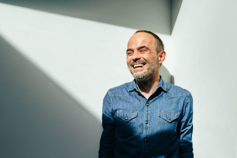 Un homme avec une barbe et une moustache se tient debout contre un mur gris clair, souriant chaleureusement les yeux fermés. Il porte une chemise en jean bleu boutonnée avec deux poches avant. La lumière du soleil projette une ombre triangulaire autour de lui, créant un effet de lumière intéressant, évoquant un sentiment d'amour inconditionnel.