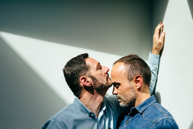 Deux hommes se tiennent côte à côte dans un moment intime d'amour inconditionnel. L'homme de gauche, aux cheveux noirs courts et à la barbe, embrasse doucement le front de l'autre homme, aux cheveux châtains courts et à la barbe, les yeux fermés. Ils sont éclairés par un faisceau de lumière triangulaire provenant d'une haute fenêtre.