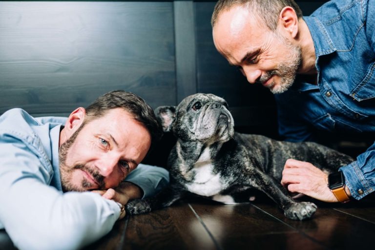 Deux hommes à la barbe taillée de près sont allongés sur un plancher en bois sombre, leurs visages proches d'un petit bouledogue français gris et tigré. L'un des hommes regarde la caméra, tandis que l'autre sourit au chien, qui lève les yeux avec une expression curieuse. Ils semblent détendus et heureux, incarnant l'amour inconditionnel.