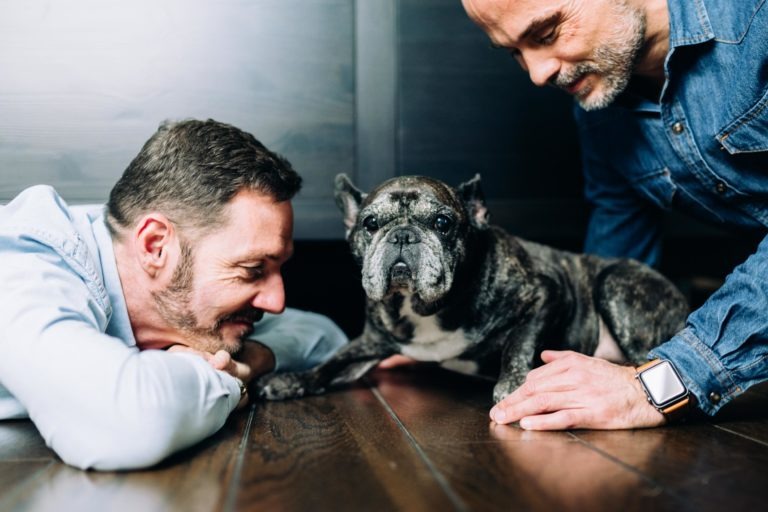 Deux hommes sourient, allongés sur un plancher en bois avec un chien tigré, incarnant un air d'amour inconditionnel. L'homme de gauche a ses mains sous son menton, tandis que l'homme de droite, portant une montre connectée, touche doucement le chien. Ils portent des chemises à manches longues décontractées sur un fond de mur sombre.