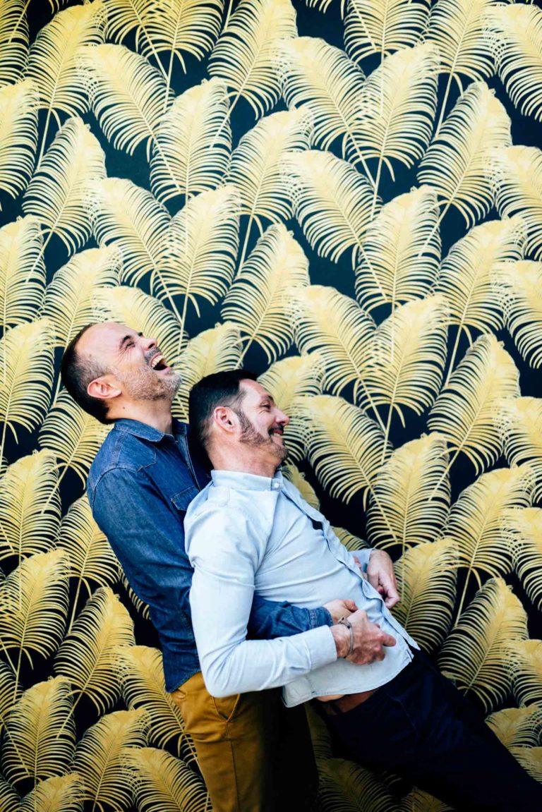 Deux hommes rient joyeusement en s'appuyant l'un sur l'autre devant un mur noir orné d'un motif de feuilles de palmier jaune vif. L'un porte une chemise bleue et un pantalon moutarde, l'autre une chemise bleu clair et un pantalon foncé, incarnant l'essence de l'amour inconditionnel.