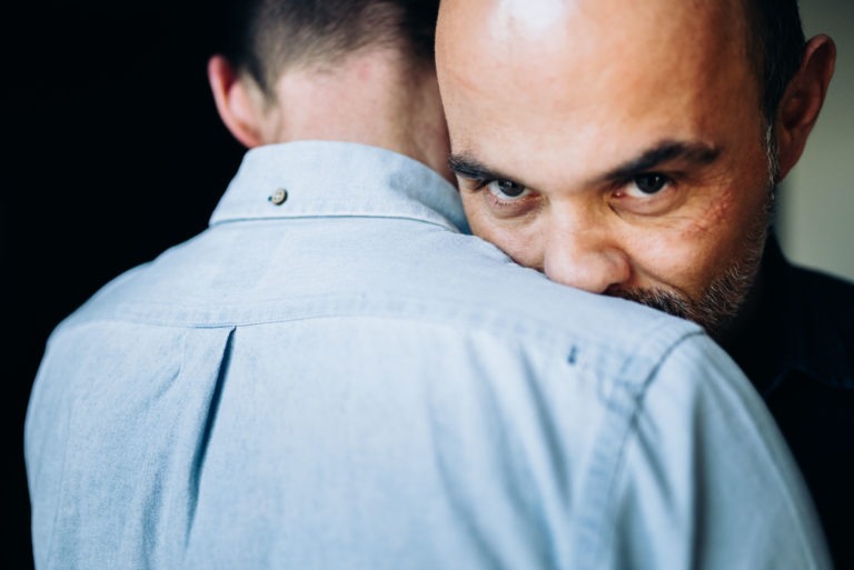 Gros plan de deux hommes qui s'embrassent. L'homme qui fait face à la caméra a le crâne rasé et une courte barbe, il regarde intensément par-dessus l'épaule de l'homme qui tourne le dos à la caméra et qui porte une chemise boutonnée bleu clair. Le fond sombre souligne l'intimité et l'amour inconditionnel de leur moment.