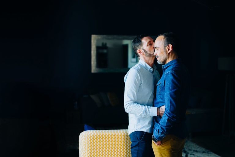 Une personne portant une chemise bleu clair embrasse doucement une autre personne portant une chemise en jean sur le front, incarnant l'amour inconditionnel. Ils se tiennent près l'un de l'autre dans une pièce faiblement éclairée aux murs sombres et à l'arrière-plan flou, qui comprend un miroir encadré. Une chaise à motifs jaunes est partiellement visible au premier plan.