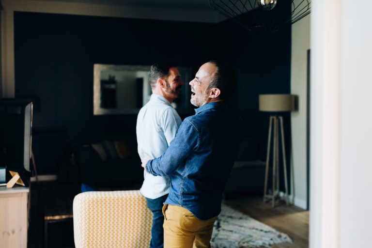 Deux hommes sont vus en train de rire et de s'embrasser dans un salon chaleureusement éclairé, rayonnant d'amour inconditionnel. L'un porte une chemise blanche et l'autre une chemise bleue avec un pantalon couleur moutarde. La pièce présente des murs bleu foncé, une lampe sur pied, un miroir au mur et une chaise et un tapis à motifs.