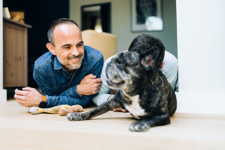 Deux hommes sont allongés sur le sol à côté d'un bouledogue français tigré, qui fait face à la caméra avec une girafe en peluche à proximité. L'homme à gauche porte une chemise en jean bleu et sourit chaleureusement au chien. Une armoire en bois et un mur en miroir sont visibles en arrière-plan, soulignant leur lien d'amour inconditionnel et créant une ambiance intérieure chaleureuse.