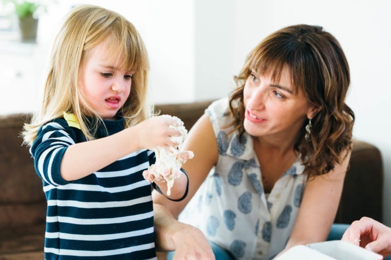 Un jeune enfant aux longs cheveux blonds, vêtu d'une chemise rayée noire et blanche, lève ses mains sales couvertes de pâte ou de bave, les regardant avec une expression curieuse. Une femme aux cheveux châtains mi-longs, vêtue d'un haut sans manches à motifs floraux, sourit en observant la réaction de l'enfant - de véritables moments simples en famille.