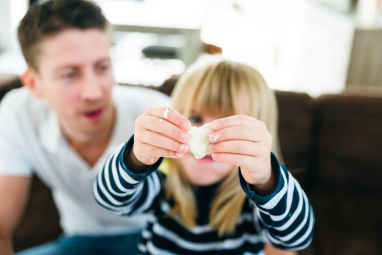 Une enfant en chemise rayée tient un morceau de pâte à modeler blanche, en se concentrant sur ses mains. Un homme en chemise blanche est assis légèrement hors champ à l'arrière-plan, la regardant. Des moments simples en famille sont partagés à l'intérieur avec une lumière vive et naturelle, suggérant une activité ludique et créative.