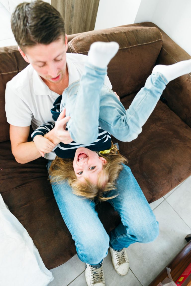 Un homme assis sur un canapé marron tient joyeusement un enfant riant à l'envers sur ses genoux. L'homme porte un polo blanc et un jean bleu clair, tandis que l'enfant, vêtu d'une chemise à rayures bleu marine et d'un jean bleu clair, lève les yeux avec joie. Le canapé et le carrelage sont visibles en arrière-plan, un instantané de moments simples en famille.