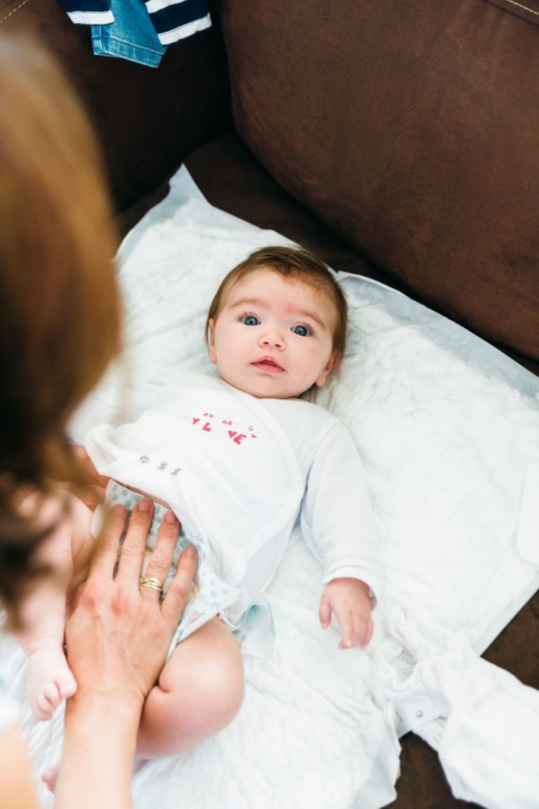 Un bébé aux grands yeux bleus est allongé sur un matelas à langer blanc, le regard tourné vers le haut. Le bébé porte une tenue blanche à petits motifs roses. On peut voir la main d'un adulte ajuster la tenue du bébé, avec un canapé marron en arrière-plan, capturant des moments simples en famille.