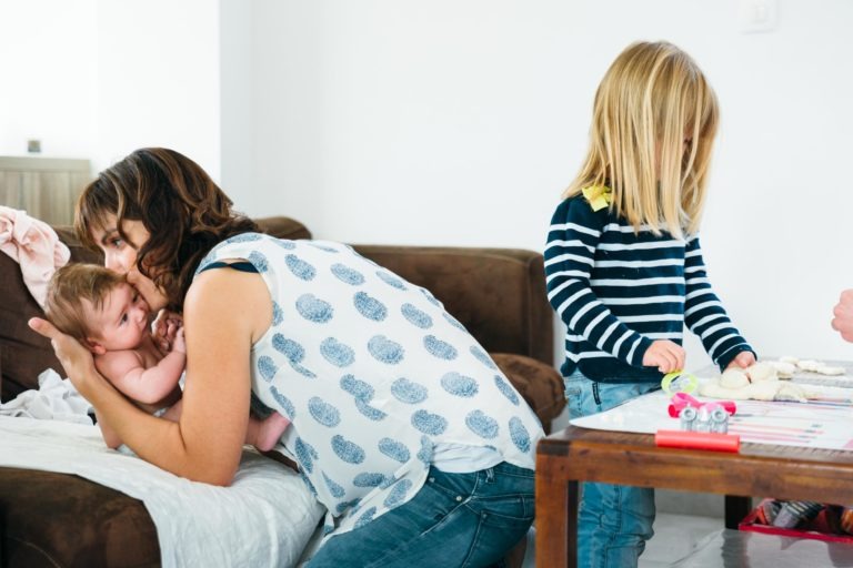 Une femme aux cheveux bruns, vêtue d'un haut sans manches à motifs, embrasse tendrement un bébé allongé sur une surface recouverte d'une couverture. Profitant des moments simples en famille, à proximité, un jeune enfant aux longs cheveux blonds, vêtu d'une chemise rayée, joue avec des jouets sur une table. L'arrière-plan présente un canapé marron et un mur blanc.