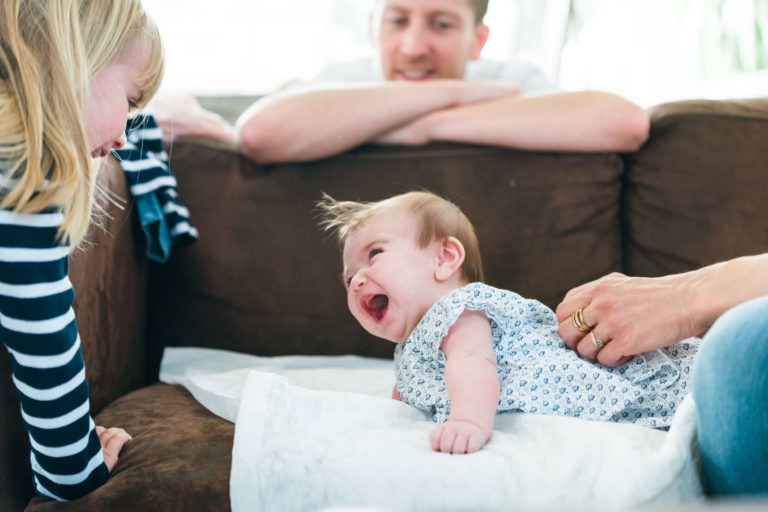 Un bébé est allongé sur un canapé et rit tandis qu'un adulte le touche de la main. Un jeune enfant aux cheveux blonds portant une chemise rayée se penche vers le bébé en souriant. Un adulte aux cheveux courts les regarde depuis l'arrière-plan, les bras tendus sur le dossier du canapé - de véritables moments simples en famille.