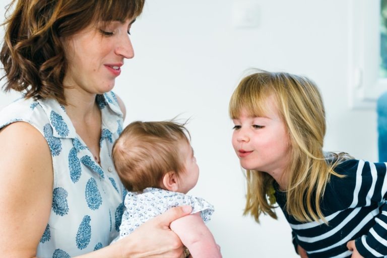 Une femme tient un bébé dans ses bras et sourit tandis qu'une jeune fille aux cheveux blonds se penche vers elle, semblant embrasser le bébé. La femme a des cheveux bruns courts et ondulés et porte un haut blanc sans manches à motifs bleus, tandis que la fille porte une chemise rayée bleu marine et blanche. Cette pièce blanche à la lumière tamisée capture de beaux moments simples en famille.