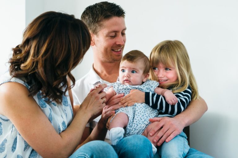 Une famille de quatre personnes est assise près les unes des autres dans ces moments simples en famille. La mère, aux cheveux bruns mi-longs, regarde avec amour le bébé dans les bras de son mari. Le père, aux cheveux bruns courts, sourit à leur jeune fille, aux cheveux blonds et portant une chemise rayée, tout en tenant le bébé.