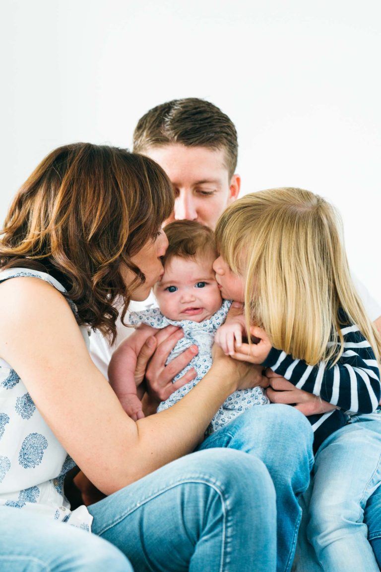 Une famille composée de deux parents et de deux enfants est représentée. La mère et l'aîné, aux cheveux blonds, embrassent un bébé sur les joues. Le père, aux cheveux bruns courts, tient le bébé dans ses bras et sourit. Ils sont assis côte à côte sur un fond blanc uni, savourant des moments simples en famille.