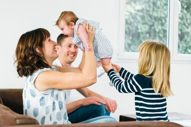 Une mère soulève un bébé dans les airs en souriant. Une jeune fille, vêtue d'une chemise rayée, tient les pieds du bébé. Un père est assis à l'arrière-plan, les regardant avec un sourire. Ils sont à l'intérieur, près d'une fenêtre avec vue sur la verdure. La scène capture des moments simples en famille pleins de joie.